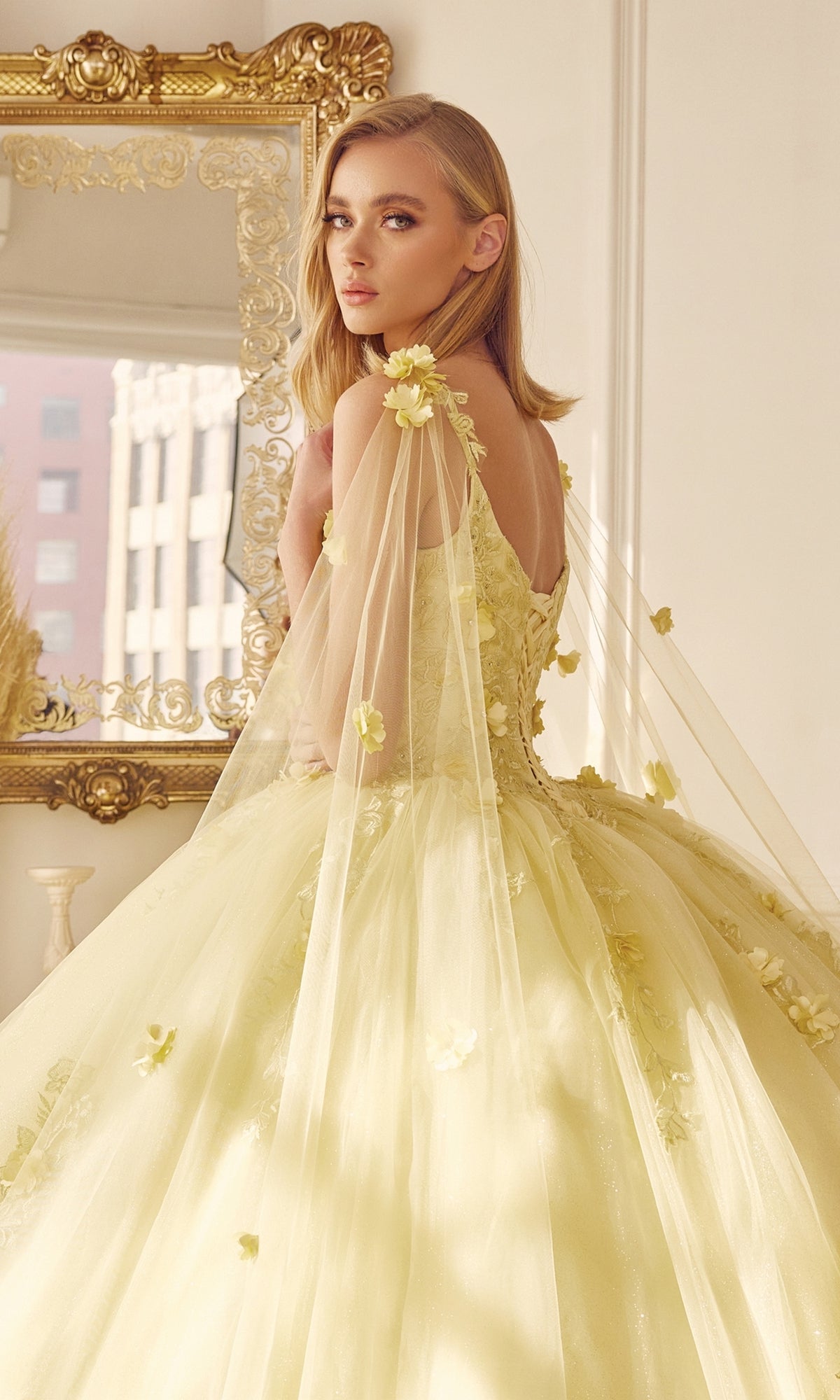 A woman wearing the Juliet 1436 Glitter Quinceañera Dress with a cape stands before a gold-framed mirror in a sunlit room.